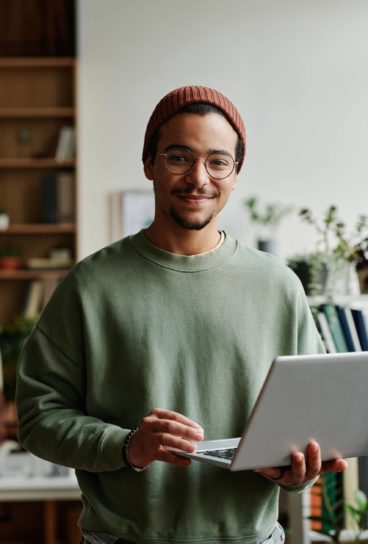 Young confident IT engineer in casualwear looking at camera in office while using laptop and working with online or coded data