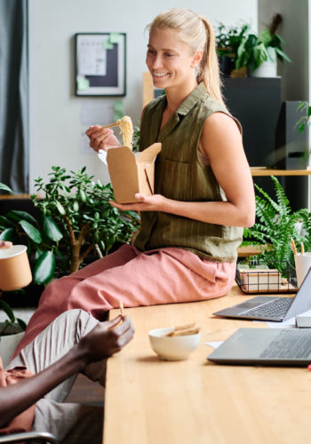 Young smiling female office manager eating wok and talking to African American male colleague sitting by workplace among green plants