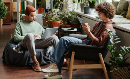 Two young serious coworkers using wireless mobile gadgets while sitting in armchairs in openspace office with variety og green plants