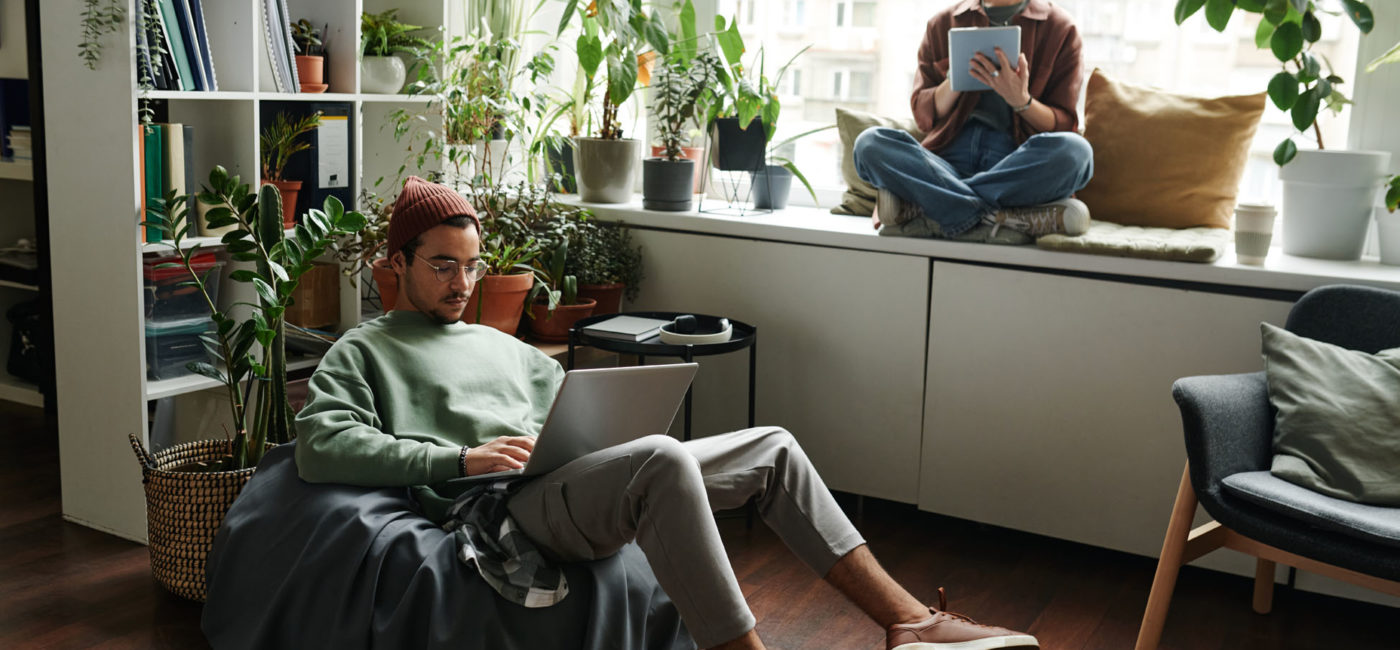 Young woman with tablet sitting on windowsill in front of male colleague with laptop networking in armchair during individual work